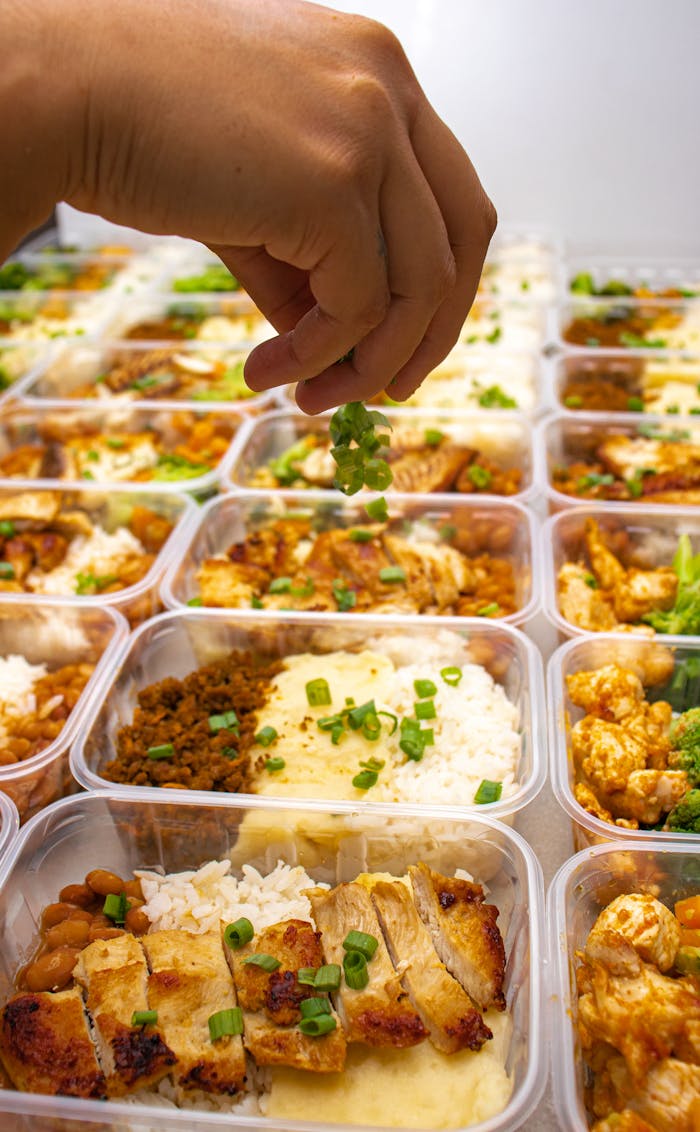 Hand sprinkling fresh herbs on meal prep containers with chicken and rice.