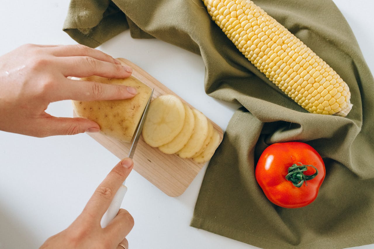 A detailed shot of hands slicing potatoes with corn and tomato on a kitchen counter.