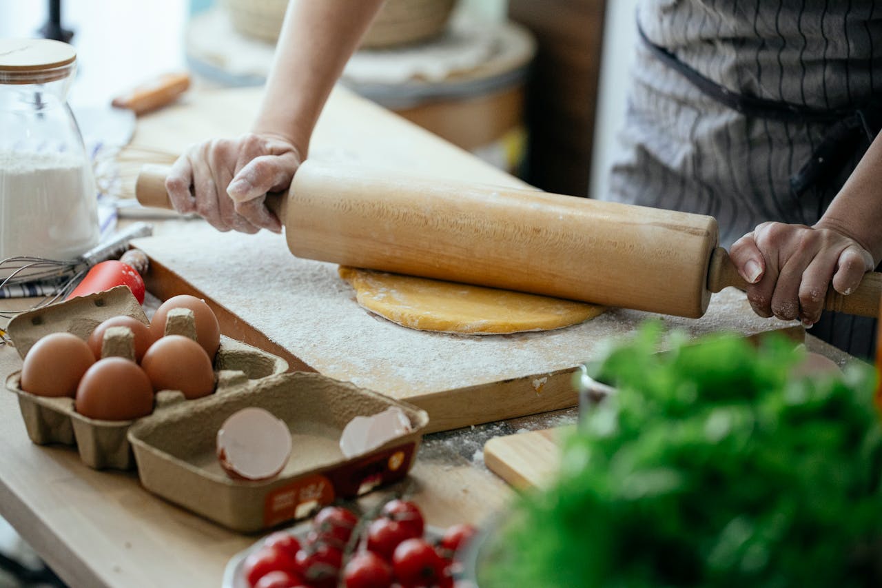 about-02 A person rolling out dough in a kitchen setting with fresh ingredients on the table.
