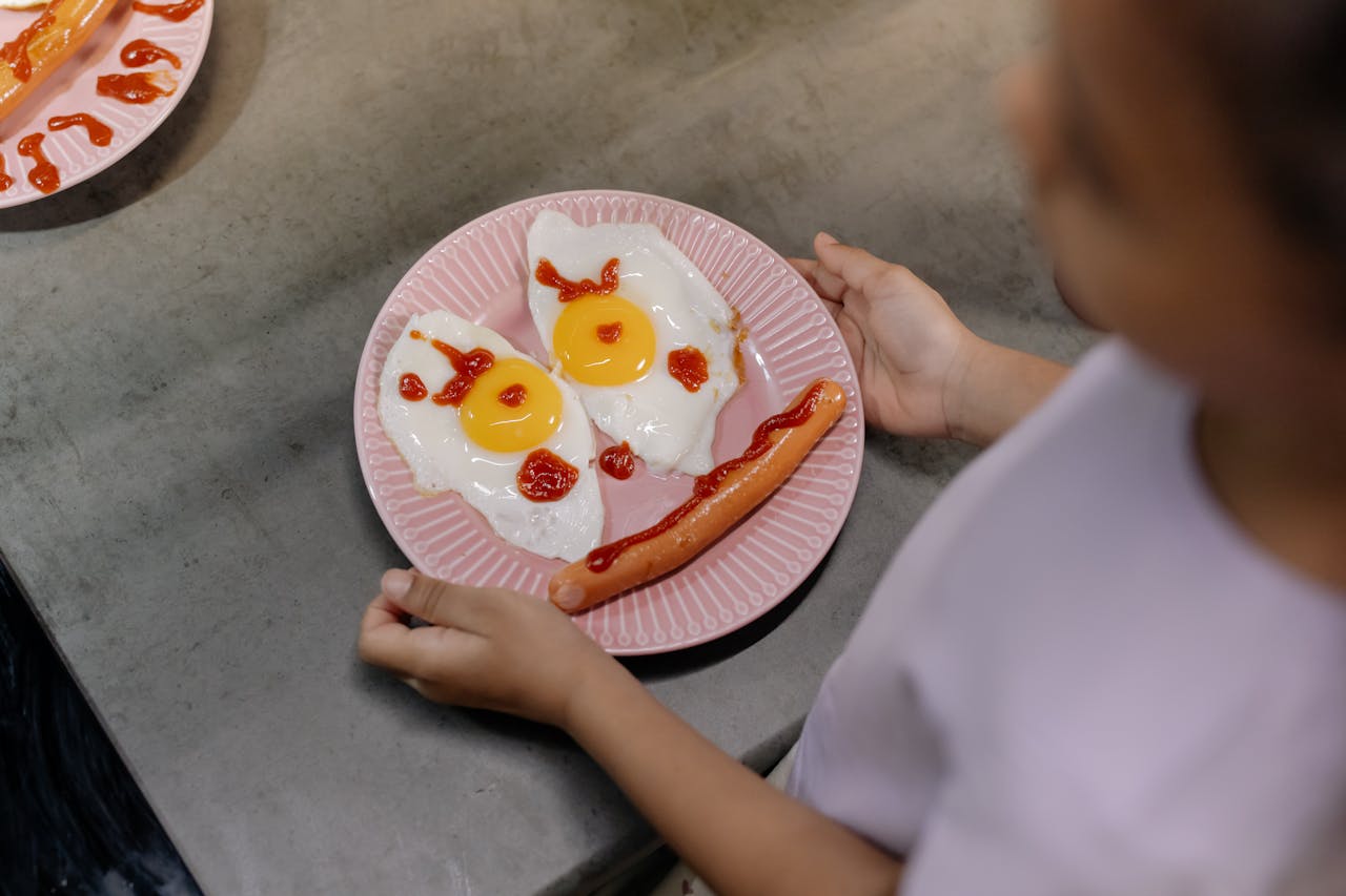 Playful breakfast setup featuring sunny-side-up eggs and sausage arranged like a smiling face on a pink plate.